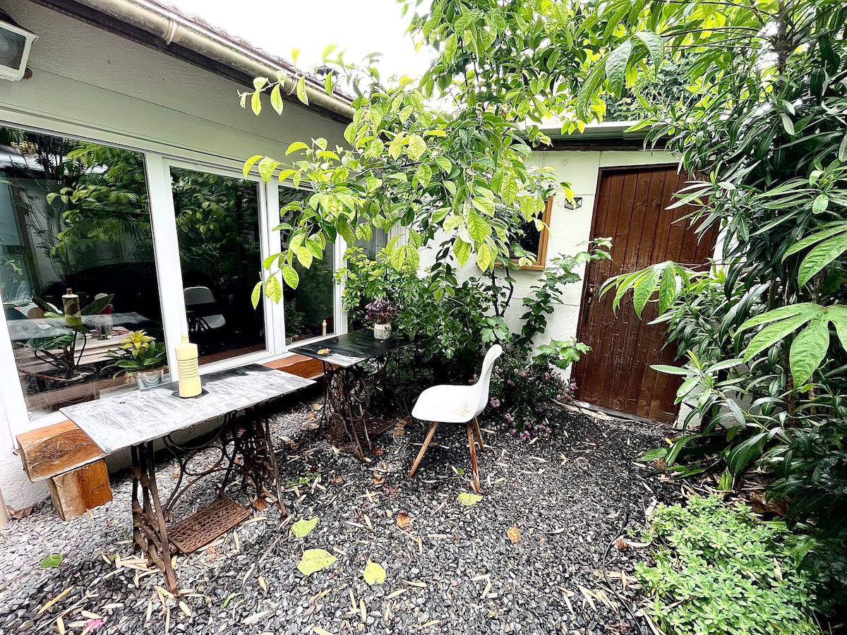 A private outdoor dining area is surrounded by lush greenery, featuring a wooden table and a white chair. Natural light filters through the large windows nearby, providing a serene spot for relaxation. The space is accented by gravel and potted plants, enhancing its tranquil ambiance.
