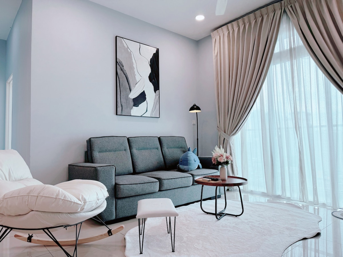 The living area features a grey sofa accompanied by a round coffee table set on a light rug. A white upholstered chair provides additional seating. Large windows adorned with curtains allow natural light to brighten the space, while a piece of abstract wall art adds a touch of style.