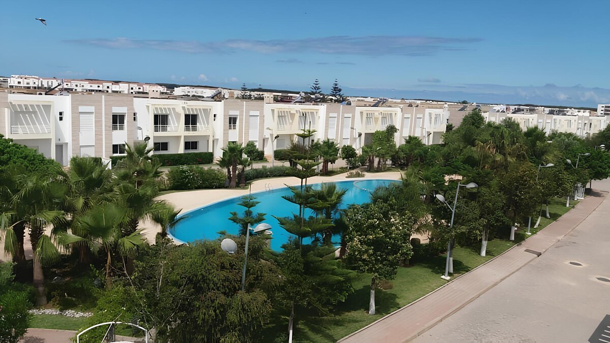 A serene view of a landscaped area featuring a large pool surrounded by palm trees and well-groomed greenery. The clean lines of the neighboring buildings are visible against a clear blue sky, contributing to the tranquil atmosphere.