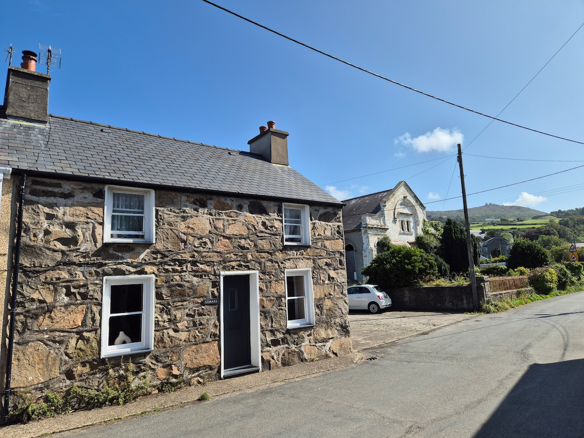 The stone cottage, characterized by its traditional architecture, features a grey slate roof and white window frames. A pathway runs alongside the exterior, leading to a nearby road. The surrounding area includes greenery and a view of hills in the background under a clear blue sky.