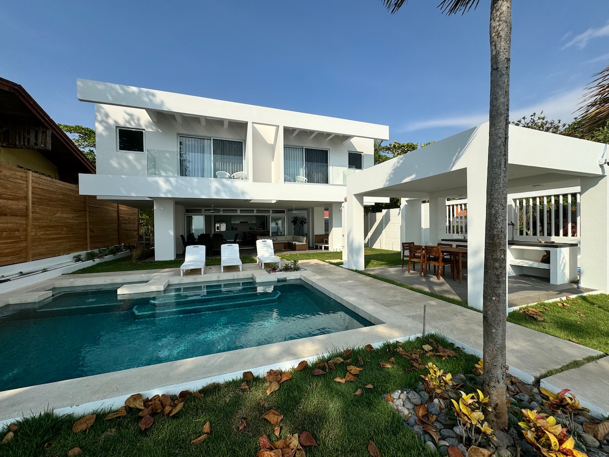 A modern two-story home is set against a clear blue sky, featuring a saltwater pool surrounded by lounge chairs. An outdoor dining area is visible under a shaded structure. Lush green grass and vibrant landscaping enhance the inviting atmosphere of the property.