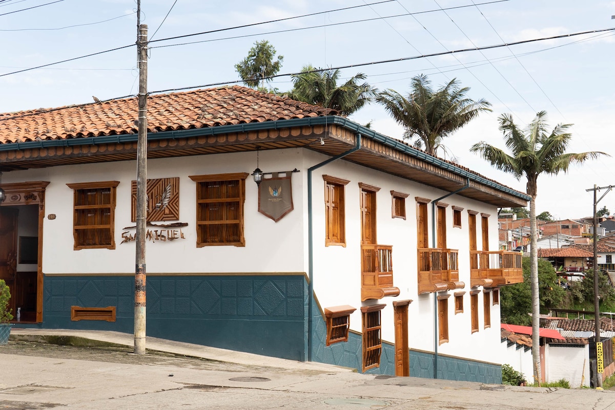 The exterior of a unique building features a combination of white and teal walls, complemented by wooden accents. The tiled roof and traditional balconies create a charming appeal. Palm trees are visible nearby, contributing to the overall distinct character of the structure.