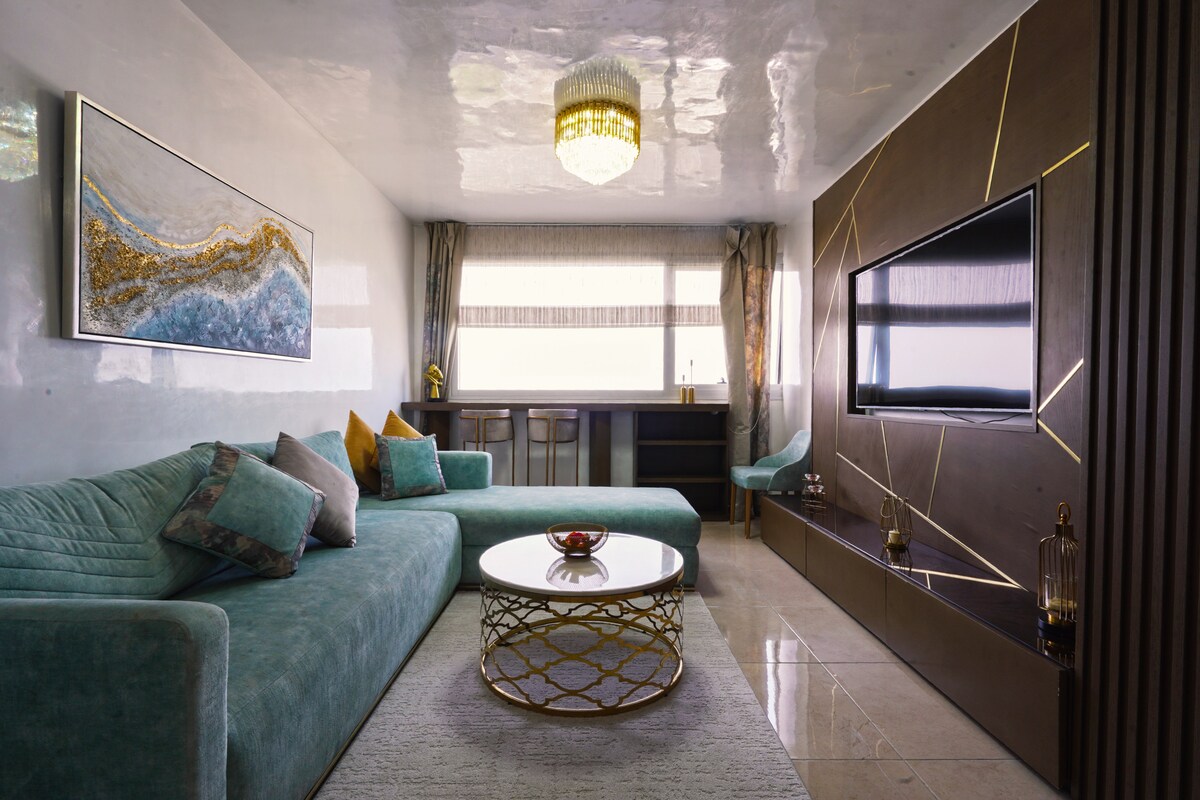 A modern living area features a large, curved turquoise sofa and a round coffee table adorned with decorative items. The space is illuminated by a chandelier above, and large windows provide natural light. A wall-mounted television complements the contemporary design.