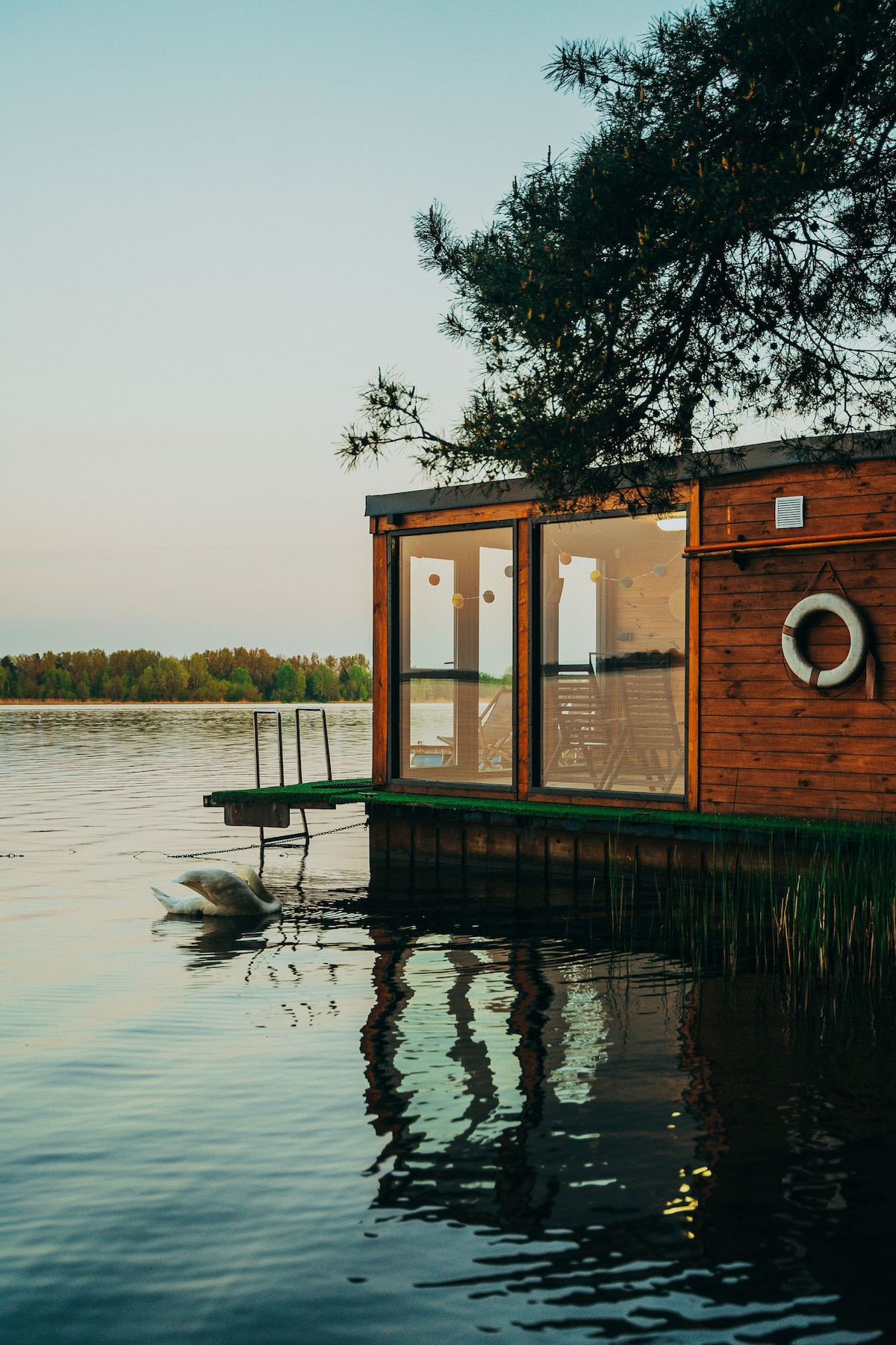 A wooden structure is positioned next to a tranquil body of water, featuring large glass windows that reflect the calm surroundings. A small dock extends into the water, with gentle ripples visible. Lush greenery borders the scene, completing a serene setting.
