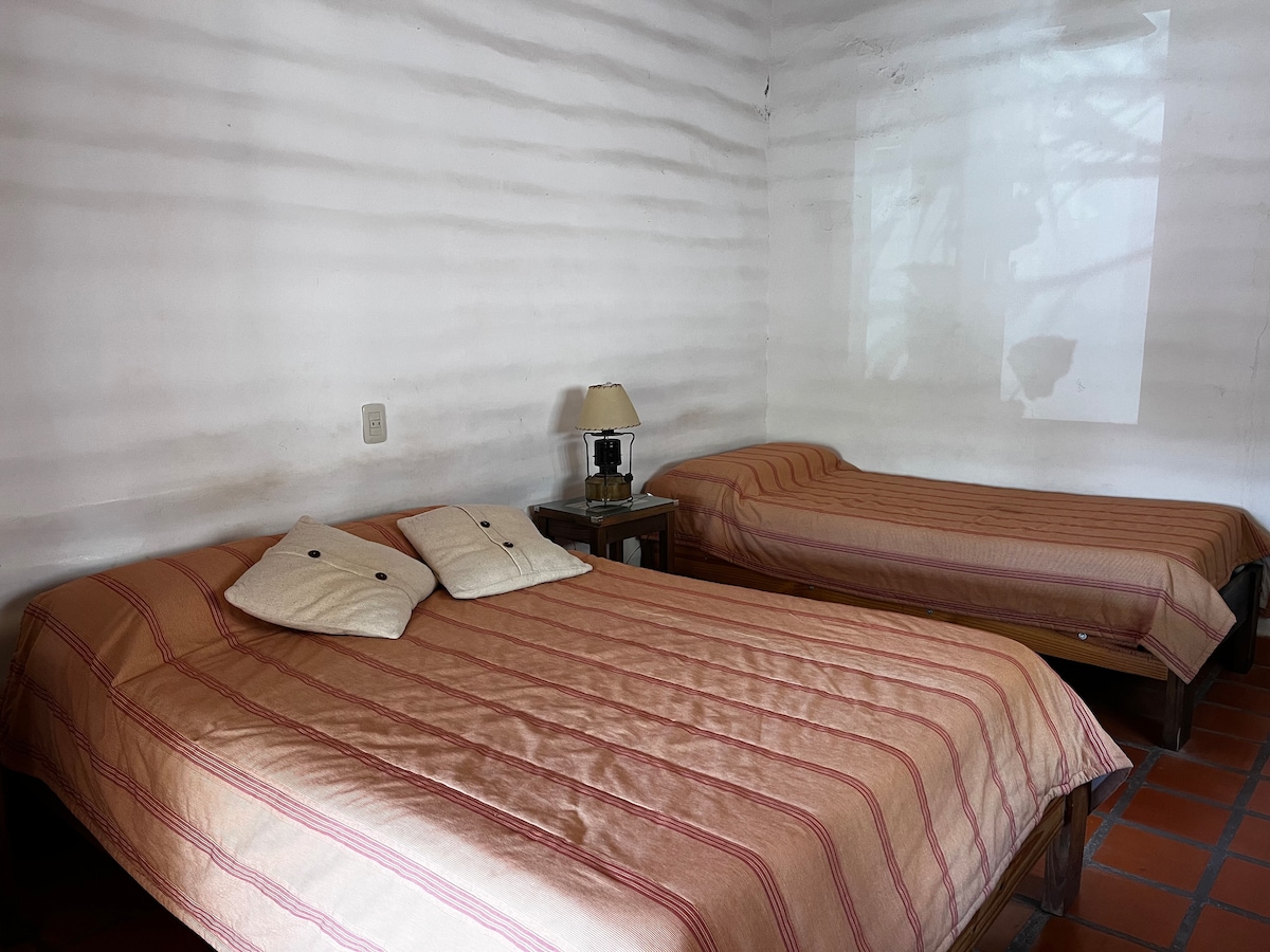 A cozy sleeping area features a double bed and a single bed, both with striped bedding. A bedside table with a lamp is positioned between the beds. The walls display a smooth, textured finish, providing a calming ambiance.