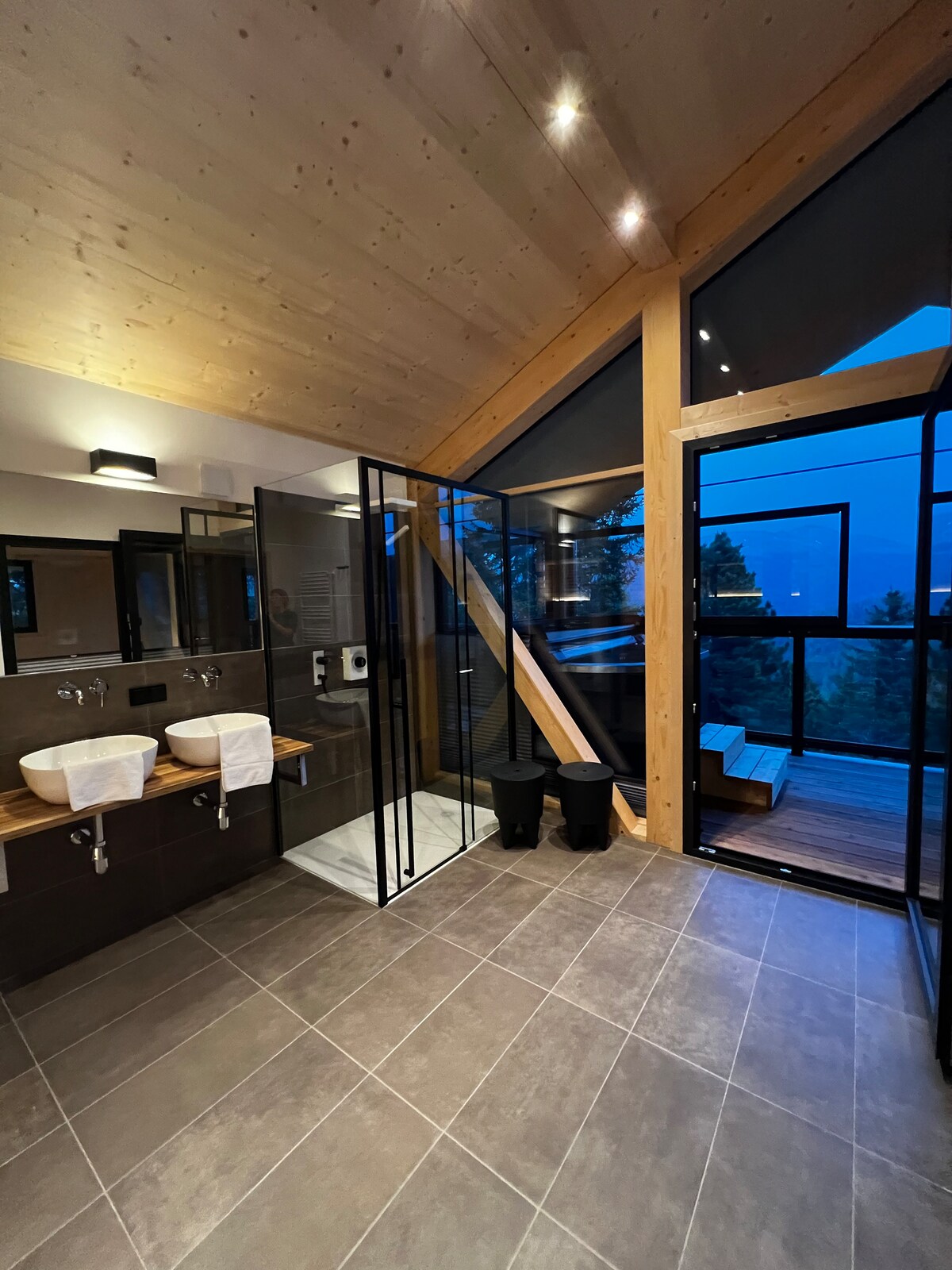 A modern bathroom is illuminated by soft lighting, featuring two sinks with sleek fixtures and a spacious walk-in shower. Panoramic windows provide a view of the outside, integrating natural scenery into the space. A private balcony area is accessible through glass doors.