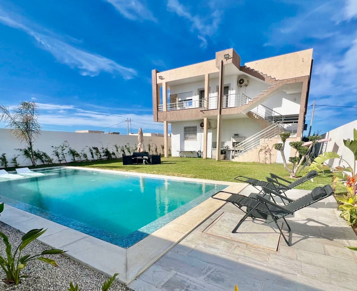 The outdoor area features a welcoming pool surrounded by lounge chairs, complemented by a well-kept lawn and plants. The two-story house showcases a modern design with balconies, inviting natural light and fresh air. Decorative elements enhance the peaceful ambiance.