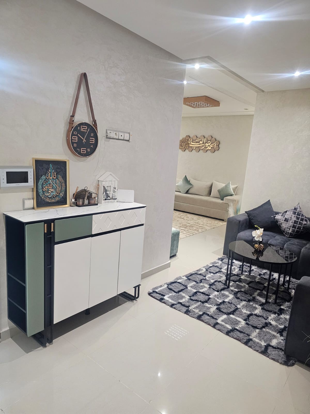 A modern living area is presented with a sleek sideboard featuring a mix of colors. A round coffee table is centered on a patterned rug, surrounded by dark sofas. Soft lighting enhances the inviting space, while decorative elements are arranged on the sideboard.