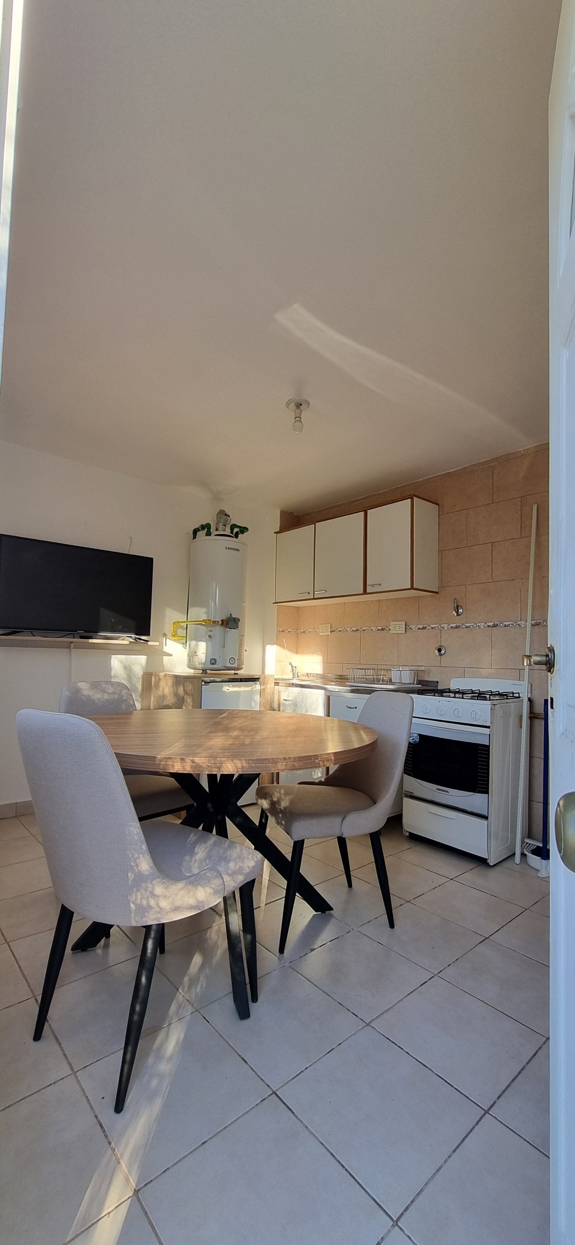 A bright kitchen area is presented with a wooden dining table surrounded by four light-colored chairs. The space includes a television mounted on the wall, minimalist cabinetry, and a stove set against a tiled backsplash, with natural light enhancing the atmosphere.