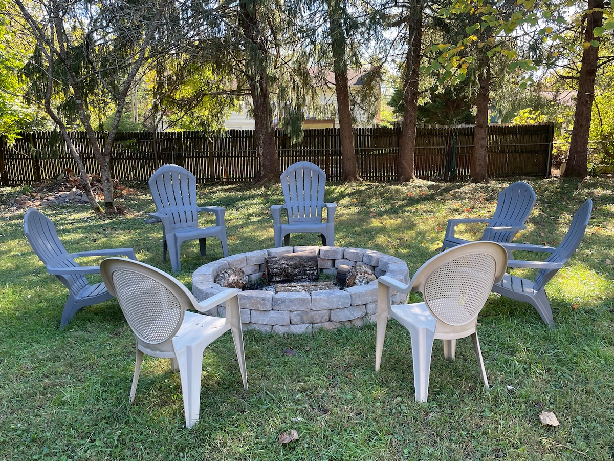 An outdoor fire pit area is surrounded by six gray adirondack chairs, arranged in a circular layout on a grassy lawn. The fire pit, constructed of stone, holds logs ready for use. A wooden fence and trees provide a natural backdrop.