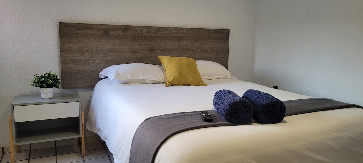 A private room features a spacious bed with a light-colored bedding set and a decorative yellow pillow. Two rolled towels are neatly placed at the foot of the bed. A small nightstand holds a potted plant, contributing to a simple and clean aesthetic.