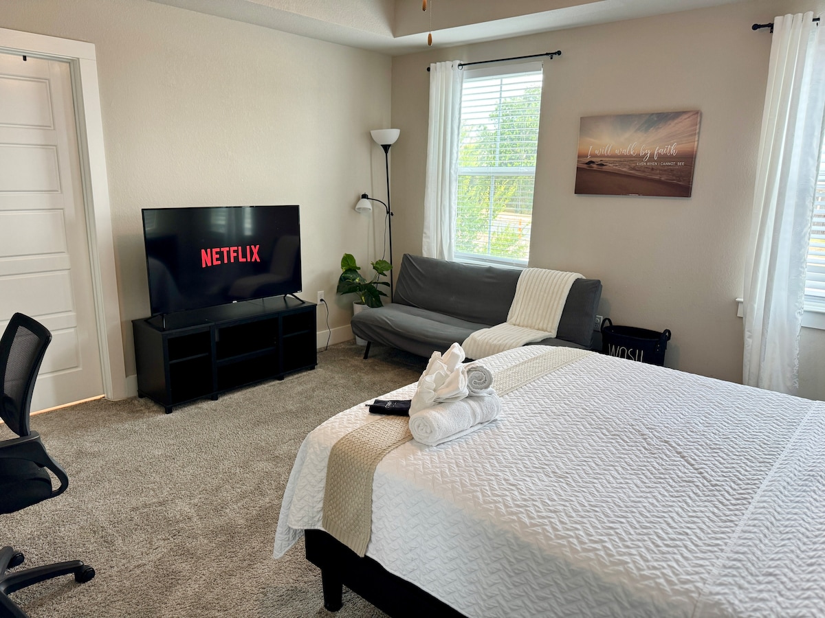A private room features a spacious bed covered with fresh linens, alongside a comfortable sofa bed. A television displaying Netflix is positioned on a black media console. Natural light enters through a window dressed with sheer curtains, and a plant adds a touch of greenery to the space.