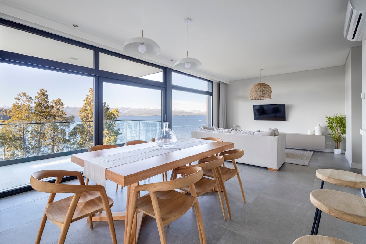 A spacious dining area features a wooden table surrounded by eight chairs, with large windows offering a view of the lake and mountains. A comfortable living area is visible, furnished with a light-colored sofa and a wall-mounted television.