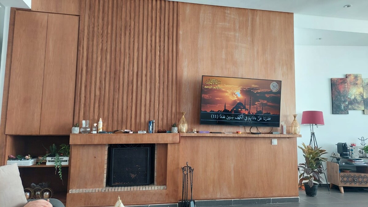 A modern living area is showcased, featuring a textured wooden accent wall with a mounted television. A minimalist fireplace with a decorative ledge is visible below the screen. Plants and soft furnishings create a cozy atmosphere, complemented by natural light filtering through adjacent spaces.