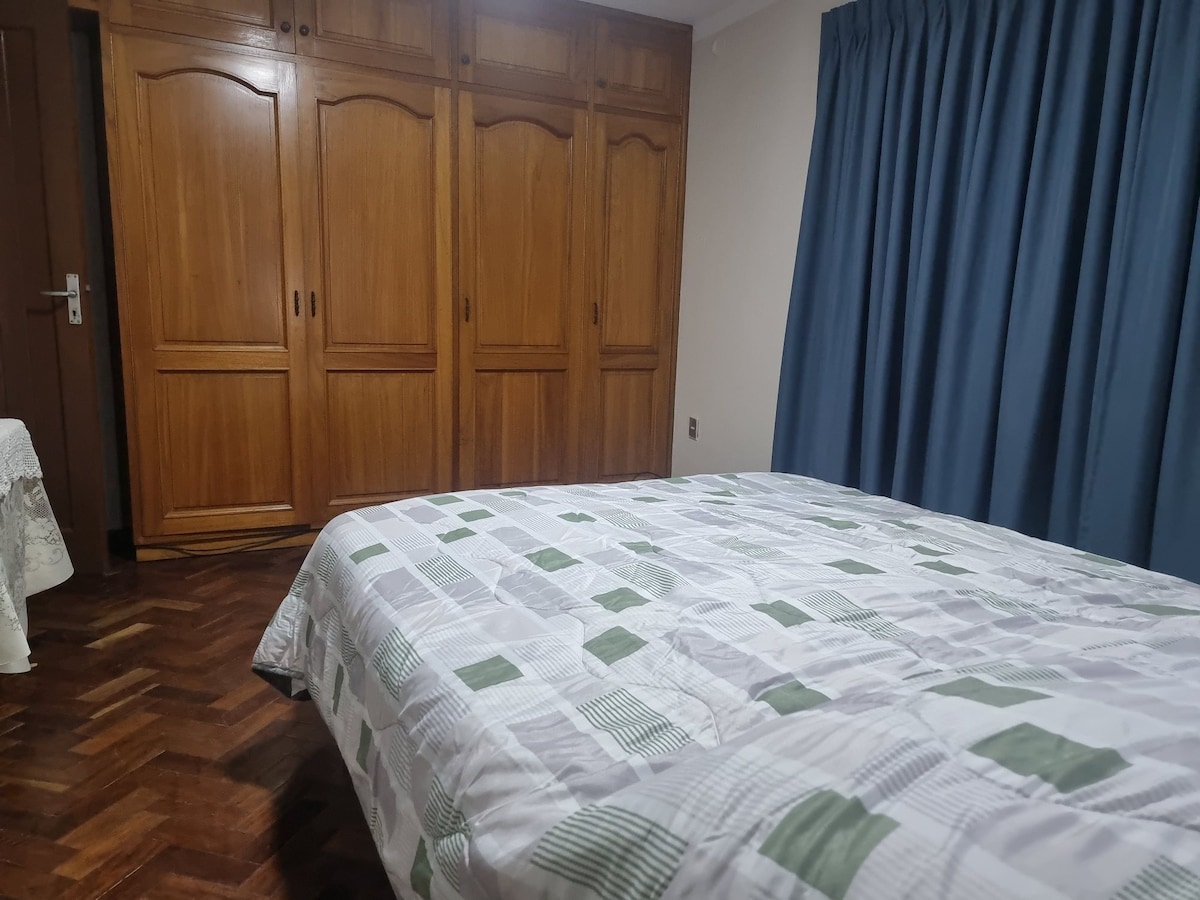 A sleeping area is equipped with a spacious bed featuring a patterned blanket. A set of wooden wardrobes occupies one wall, providing ample storage space. The room is complemented by dark blue curtains that frame a window and a small table visible in the corner.