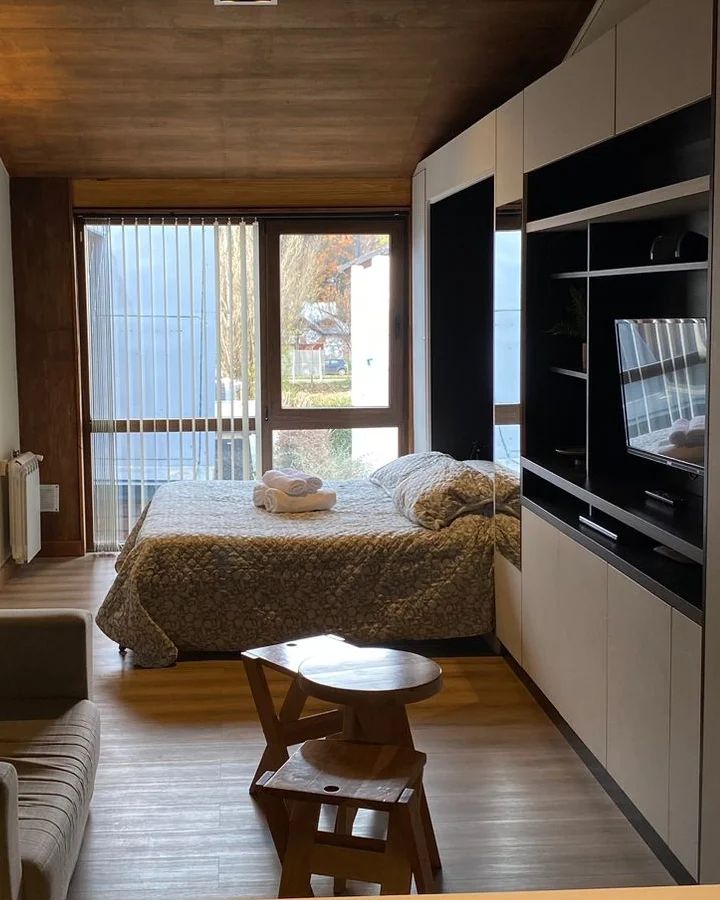 A cozy living area features a spacious bed covered with a patterned blanket. Natural light enters through large windows with blinds. A compact seating area with a small table is visible, along with a mounted television and storage unit built into the wall.