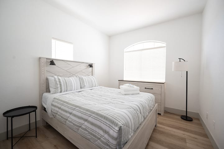 Спокојна спална соба со уредно направен кревет. Меката постелнина со риги и стилската подна ламба создаваат пријатна атмосфера, совршена за опуштање.