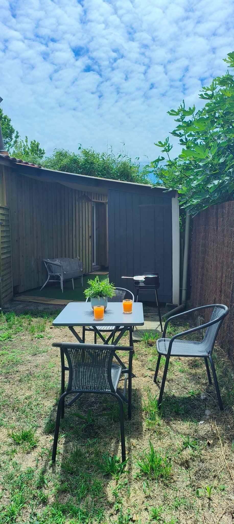 An outdoor garden space is presented with a small table and two chairs, accompanied by a barbecue. Refreshing orange drinks are placed on the table. The area is surrounded by greenery, with a shed visible in the background, and a cozy atmosphere is suggested by the setting.