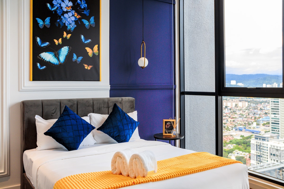 A bedroom is presented with a plush bed adorned in crisp white linens and a bright orange throw. Vibrant butterfly artwork decorates the wall, complemented by blue accent pillows. A small side table holds a framed picture, and large windows offer expansive city views.