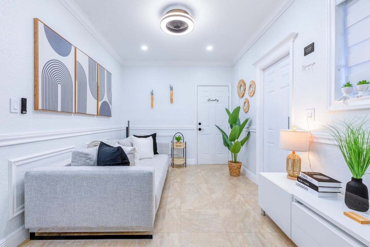 A modern living space features a light grey sectional sofa adorned with black and cream cushions. Three geometric wall art pieces hang above the couch. A potted plant and a lamp provide subtle greenery and warmth, while the room is illuminated by a stylish ceiling fixture.