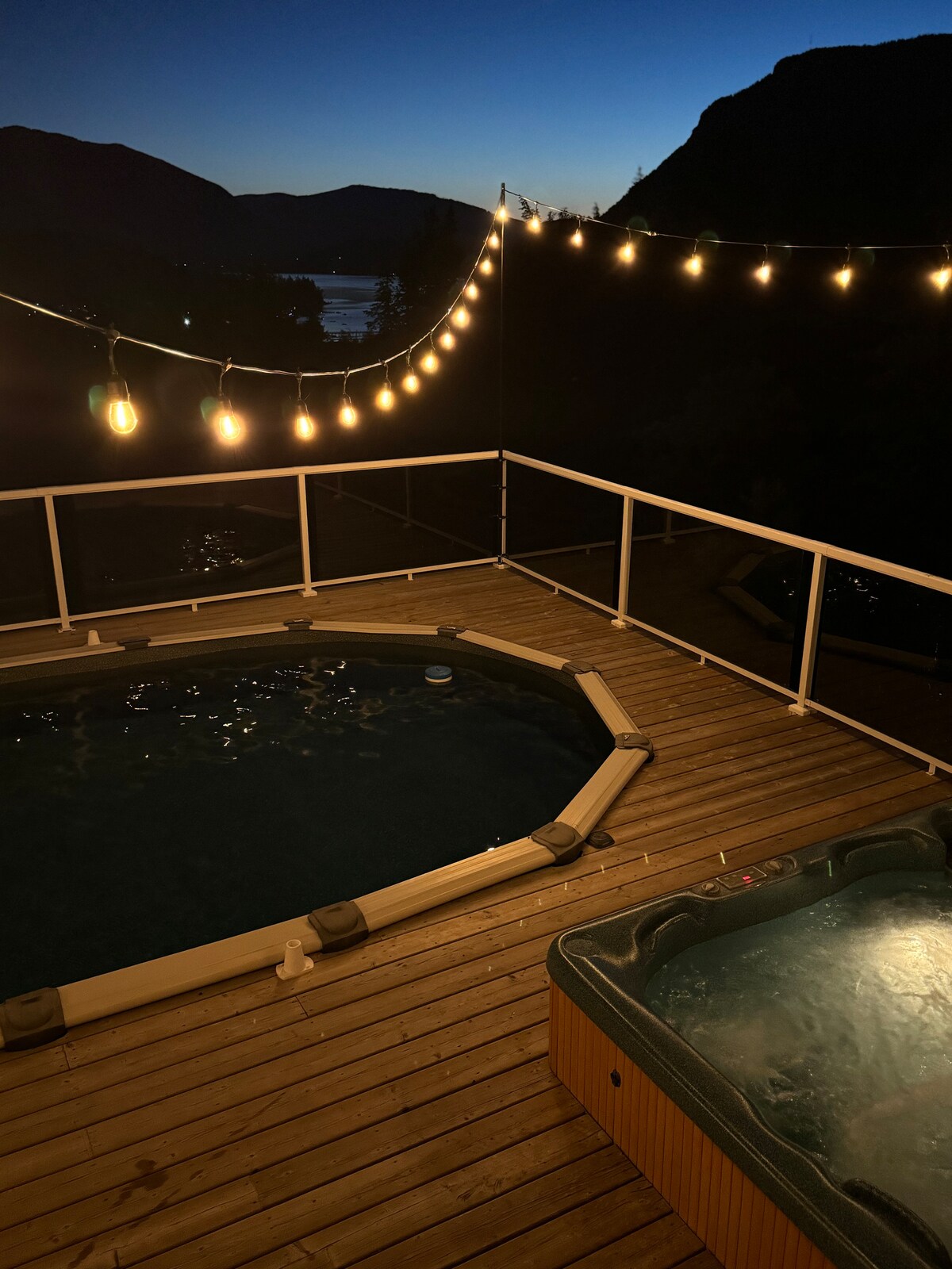 The image captures a heated pool and hot tub on a wooden deck, illuminated by string lights. The serene lake can be seen in the background, and the outline of mountains is subtly highlighted by the twilight sky.
