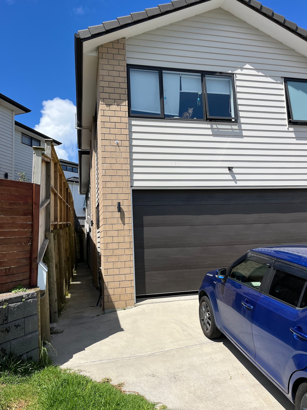 A modern two-story home is visible, showcasing a light-colored exterior with a combination of brick and cladding. A garage door is situated prominently, with a blue car parked on the driveway. Sunlight reflects off the clean surfaces, creating a bright atmosphere.