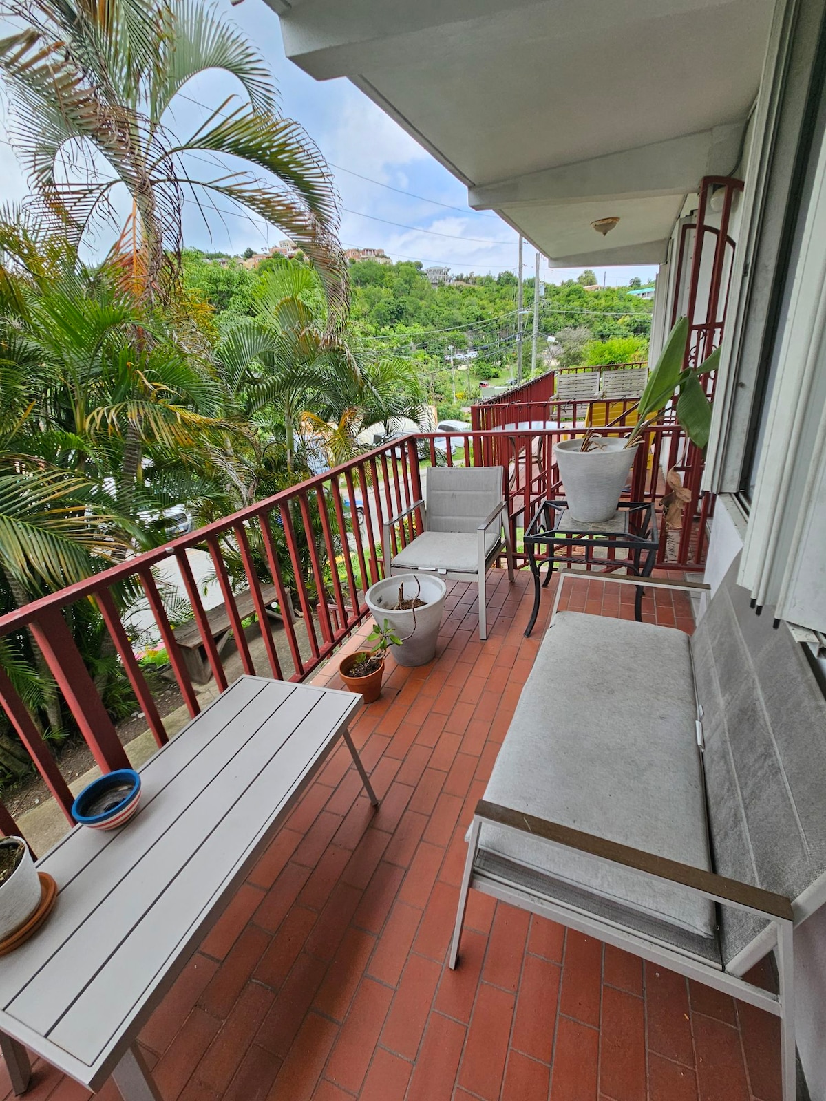 An outdoor balcony is furnished with two chairs and a small table, surrounded by lush greenery and palm trees. Several potted plants can be seen, with sunlight filtering through the foliage, creating a serene spot for relaxation.