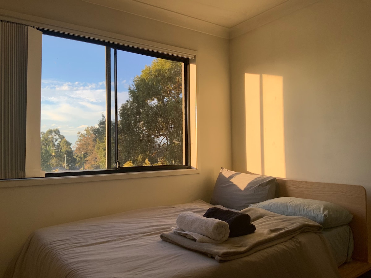 A well-lit sleeping area features a spacious bed with neatly arranged blankets and towels. Large windows allow natural light to fill the room, providing views of greenery outside. Soft shadows create a calming atmosphere during the late afternoon.