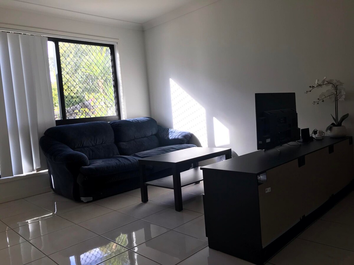 A bright living area features a dark blue couch positioned beside a low coffee table. A sleek entertainment unit is placed across from the couch, with a television set atop. Vertical blinds filter natural light from the window, creating a calm ambiance.