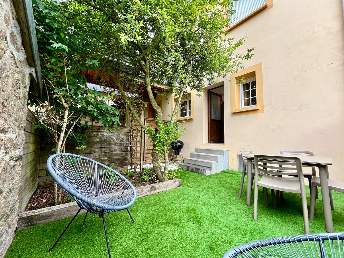 An outdoor space features artificial grass and a mix of greenery, providing a relaxing atmosphere. A small table and chairs are positioned beside a charcoal grill. The entrance to the apartment is visible, framed by charming plants and trees.