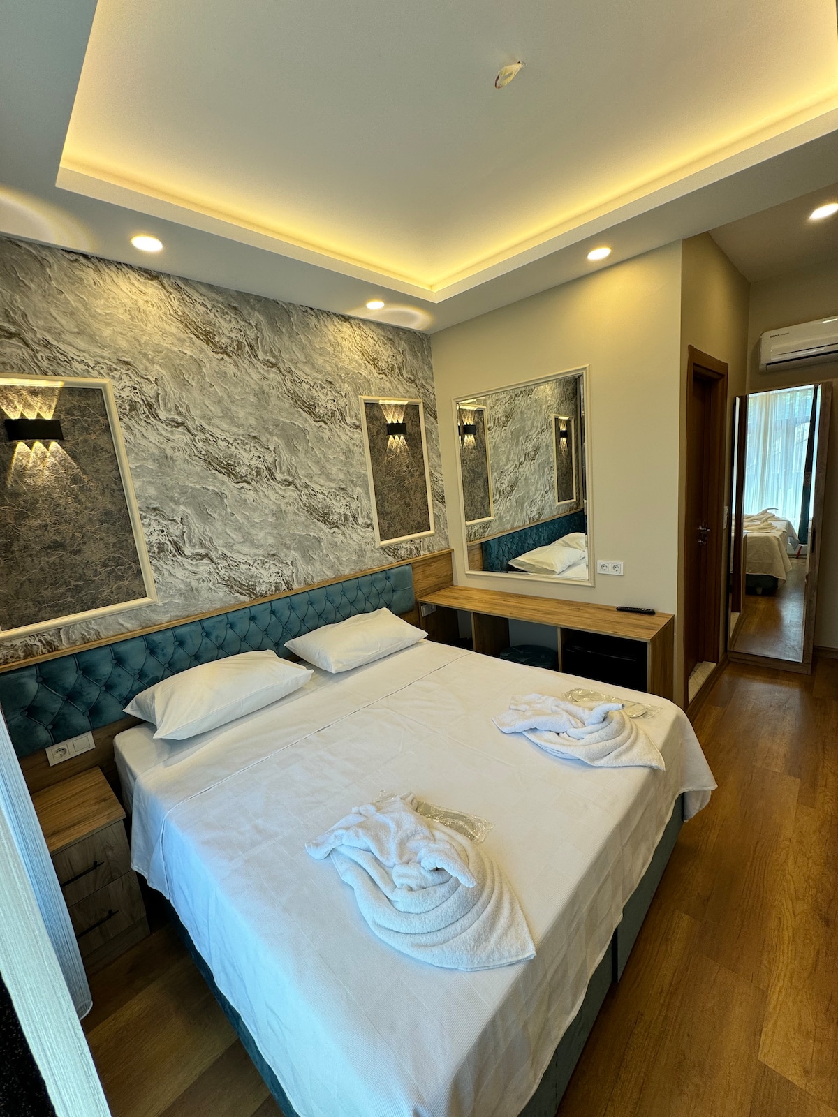 A well-appointed bedroom presents a spacious bed with neatly arranged towels. The elegant wall features a marbled design, complemented by soft lighting fixtures. A mirrored wall reflects the room's layout, while a large window allows natural light to fill the space, enhancing the inviting ambiance.