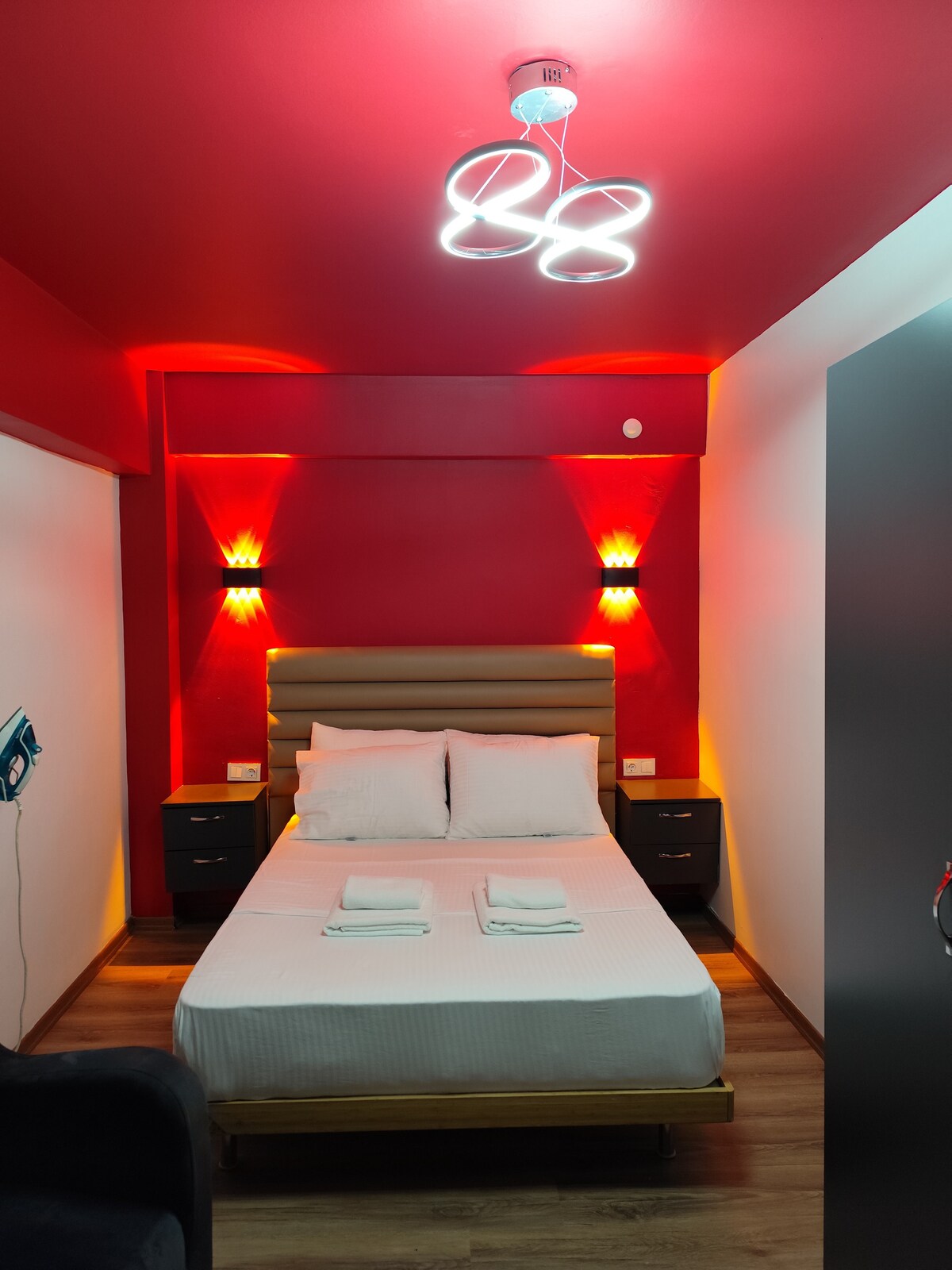 A modern sleeping area features a neatly made bed with white linens and two folded towels placed on top. The walls are painted in a bold red, complemented by stylish lighting fixtures. Dark nightstands are positioned on either side of the bed.