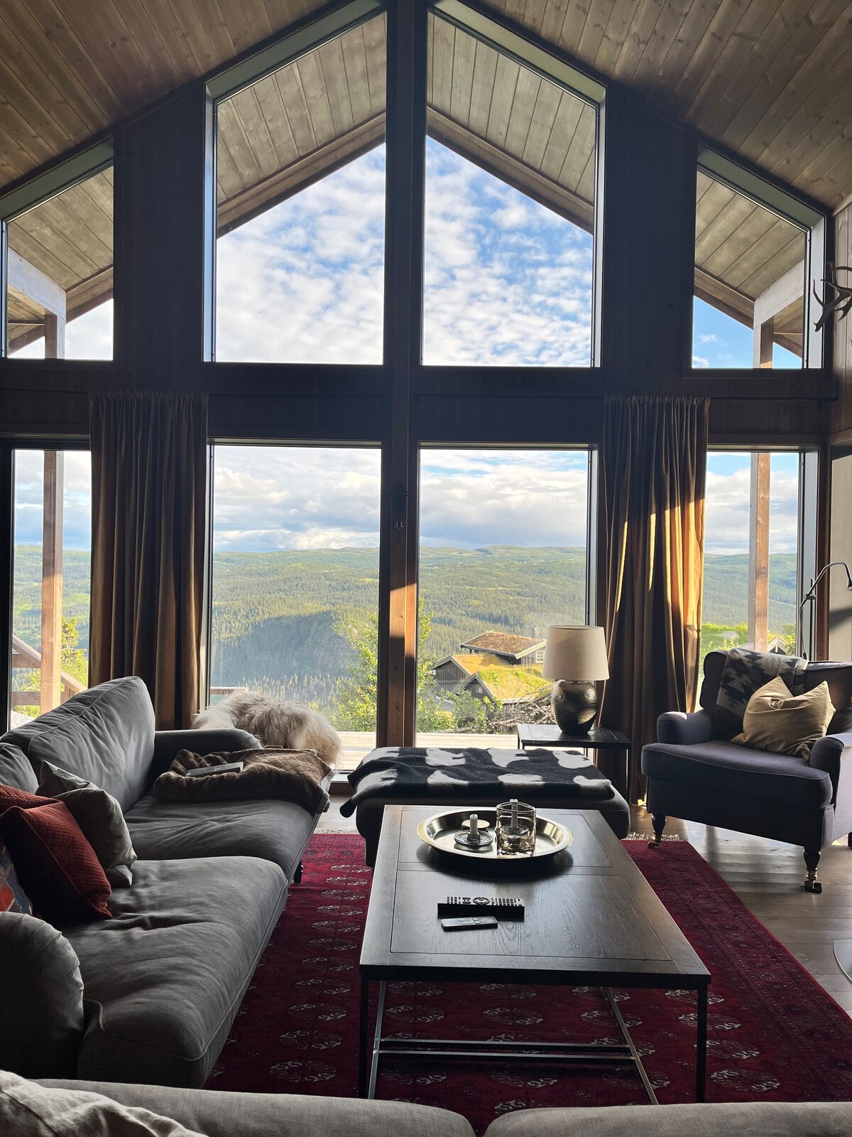 A spacious living area features large, triangular windows offering a panoramic view of the surrounding landscape. Comfortable seating includes a dark sofa adorned with pillows and a cozy armchair. A coffee table is positioned in front, accompanied by decorative items, creating an inviting space.