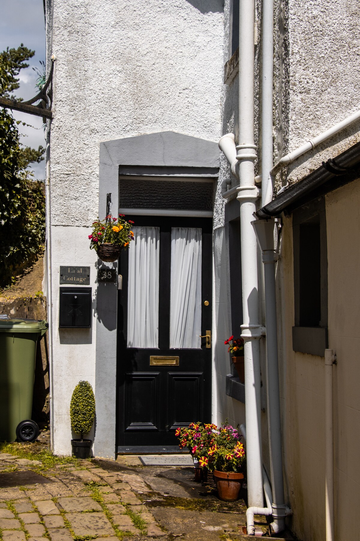 The entrance to La'al Cottage features a black door framed by potted plants, including vibrant flowers. A small decorative planter stands beside the door, and a plaque indicating the cottage's name is mounted nearby. The cobblestone pathway leads to the welcoming entrance.