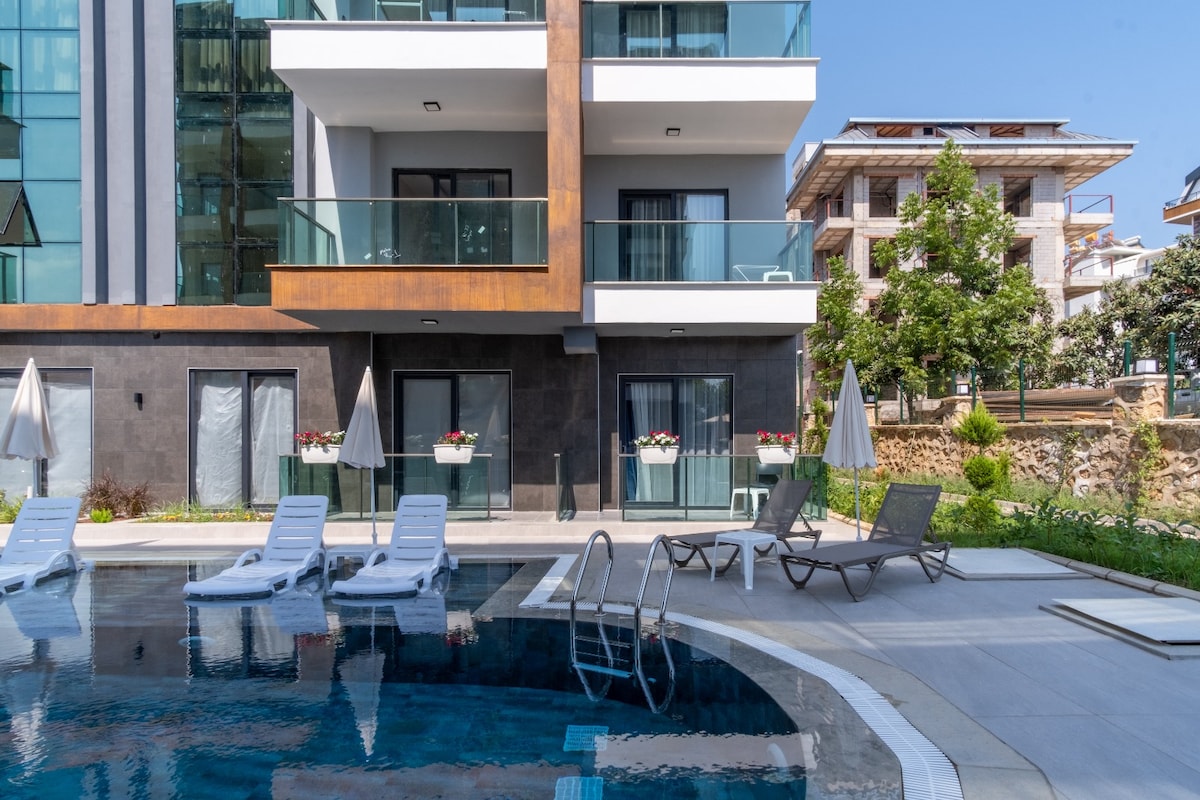 The image captures a modern building with multiple stories, featuring large glass balconies. A pool with sleek lounge chairs is visible in the foreground, accompanied by shaded umbrellas. Lush greenery is present around the property, enhancing the inviting outdoor space.