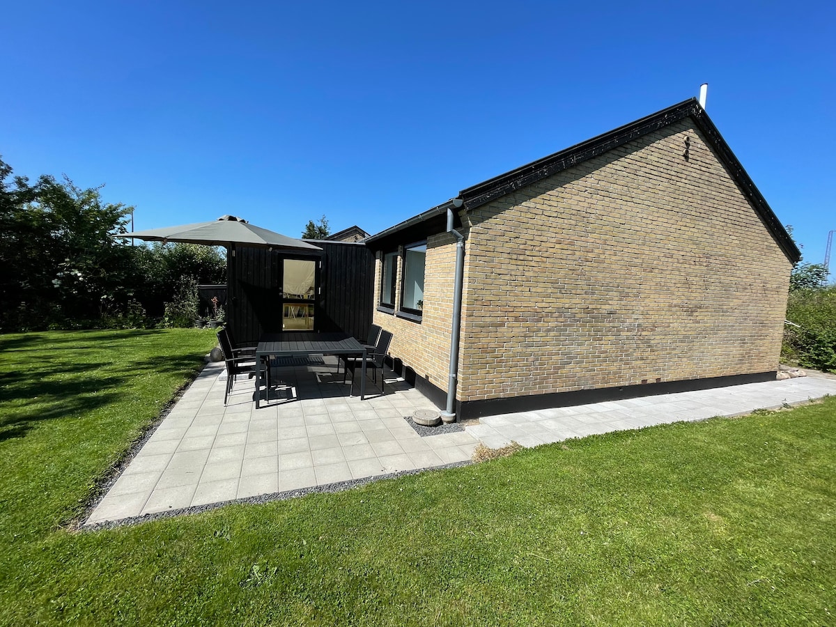 A spacious patio area is visible beside the house, featuring a large outdoor table and chairs, sheltered by a sunshade. The surrounding yard is well-maintained, with green grass and plants enhancing the outdoor space. Clear blue skies create a bright and inviting atmosphere.