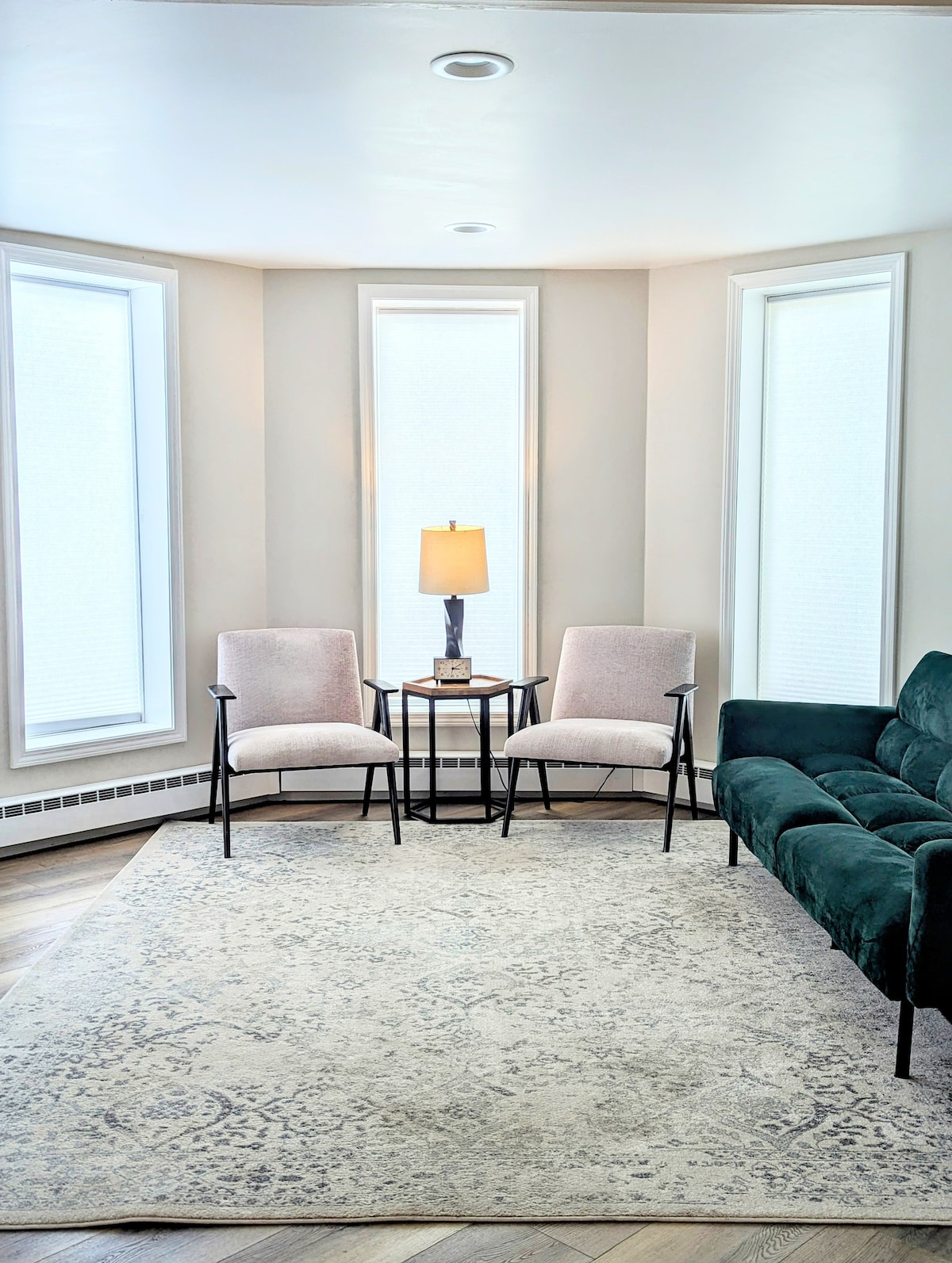 A well-lit living area features two upholstered chairs and a small side table with a lamp, positioned near large windows that allow natural light to fill the space. A plush, dark green sofa complements the neutral decor, resting atop a softly patterned area rug.