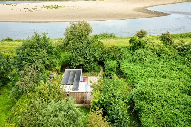 An aerial view displays a tiny house nestled among lush greenery, with a private terrace visible. The proximity to the Maas river is evident, while the surrounding nature offers a serene and secluded environment.