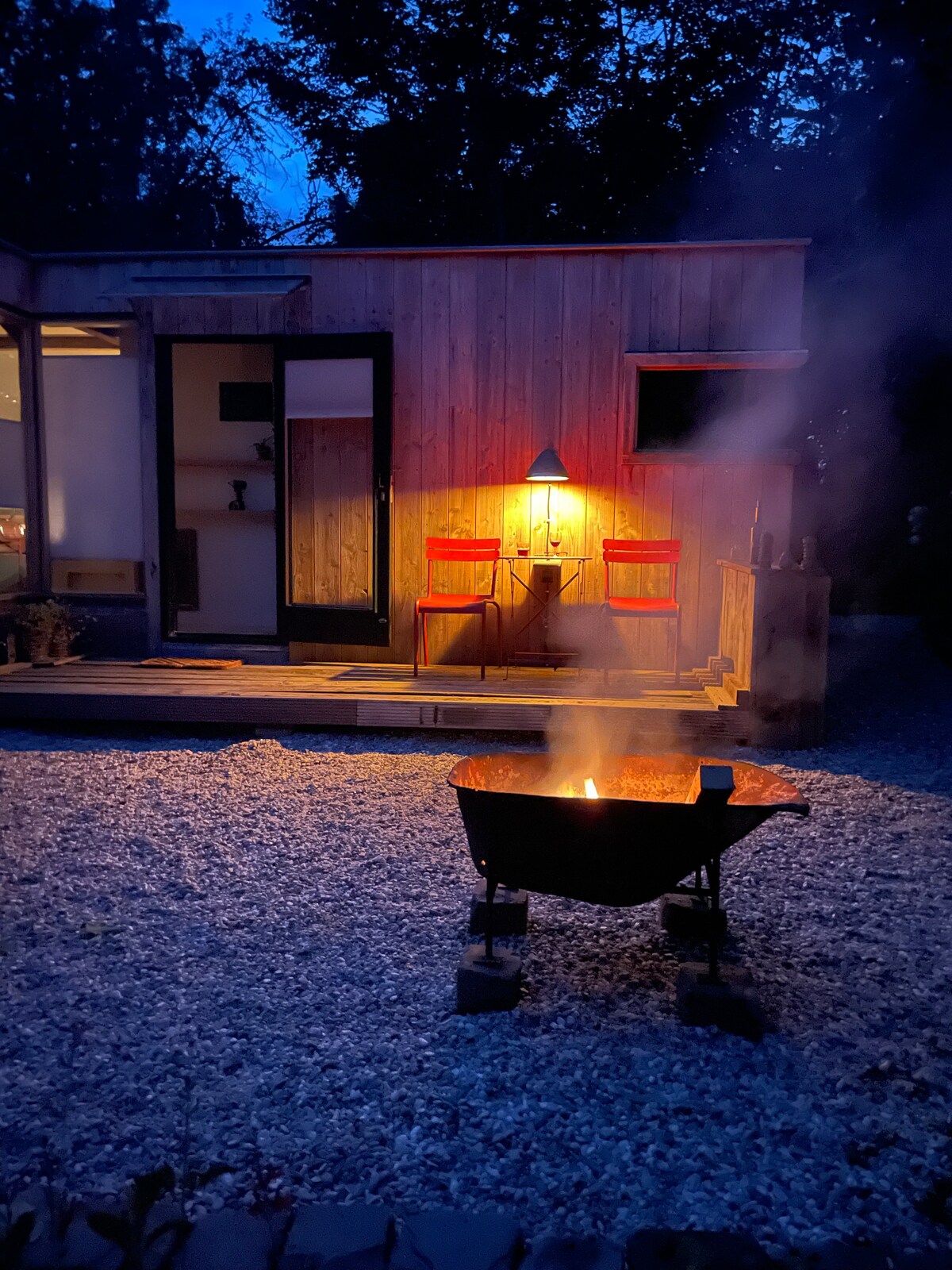 A private terrace is illuminated by a warm light from a nearby entrance, featuring two chairs positioned invitingly. A fire pit emits a gentle smoke, surrounded by gravel, creating a peaceful outdoor setting at dusk.