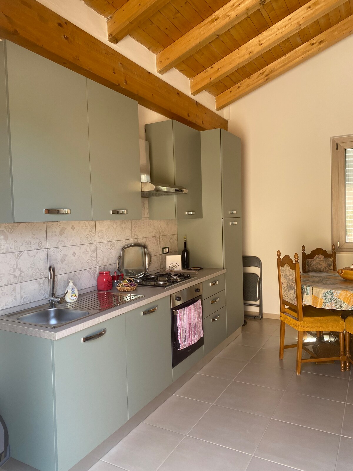 The kitchen area features a modern design with light green cabinetry and a stainless-steel stove. A dining table with wooden chairs is positioned nearby, while ample natural light is provided by a window with horizontal blinds. Exposed wooden beams add warmth to the space.