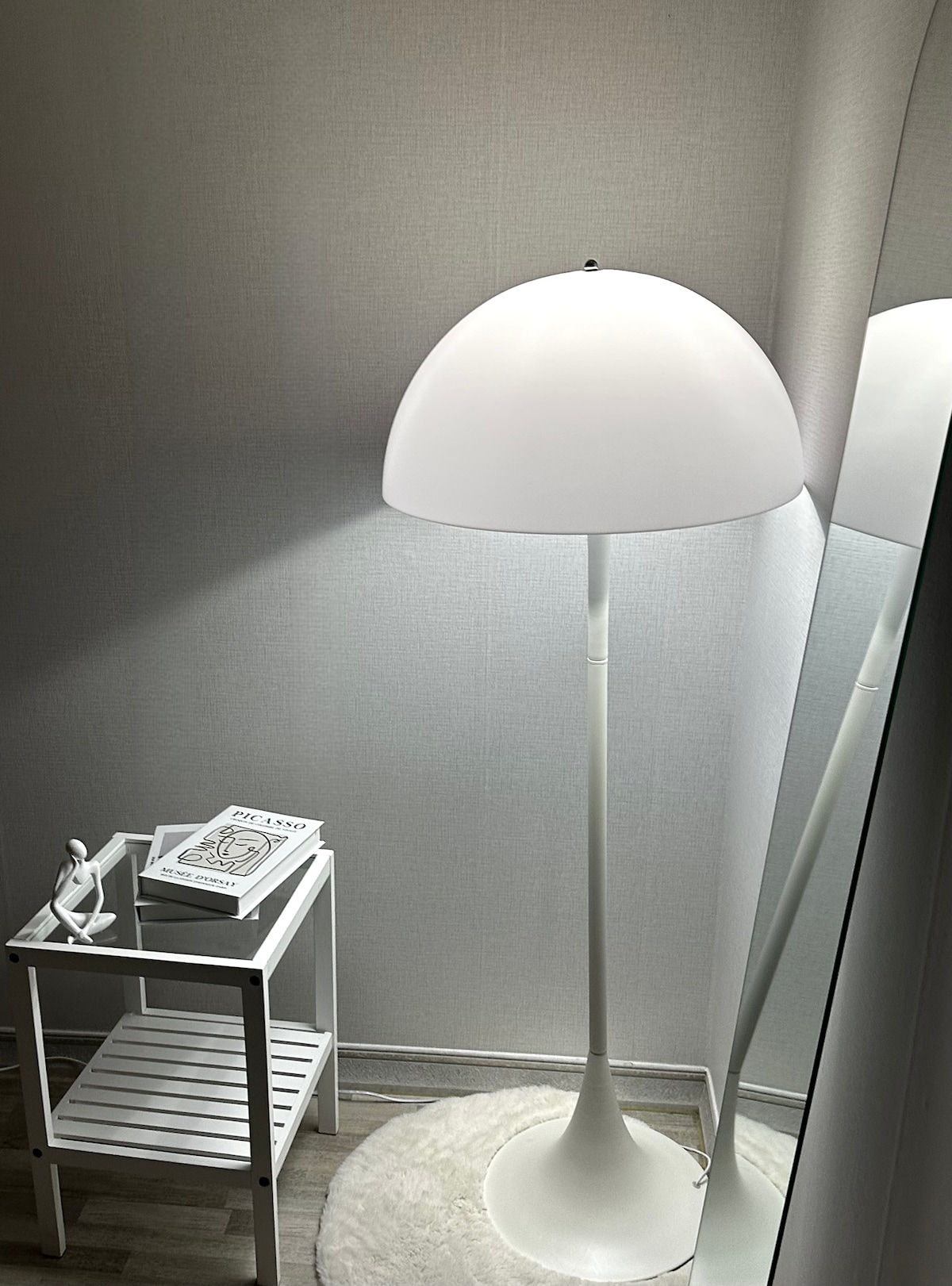 A sleek white floor lamp provides soft illumination beside a minimalist side table, which holds a book and a decorative figure. A round, plush rug is placed on the floor. The reflection of the lamp and table can be seen in a nearby mirror, enhancing the light and space.