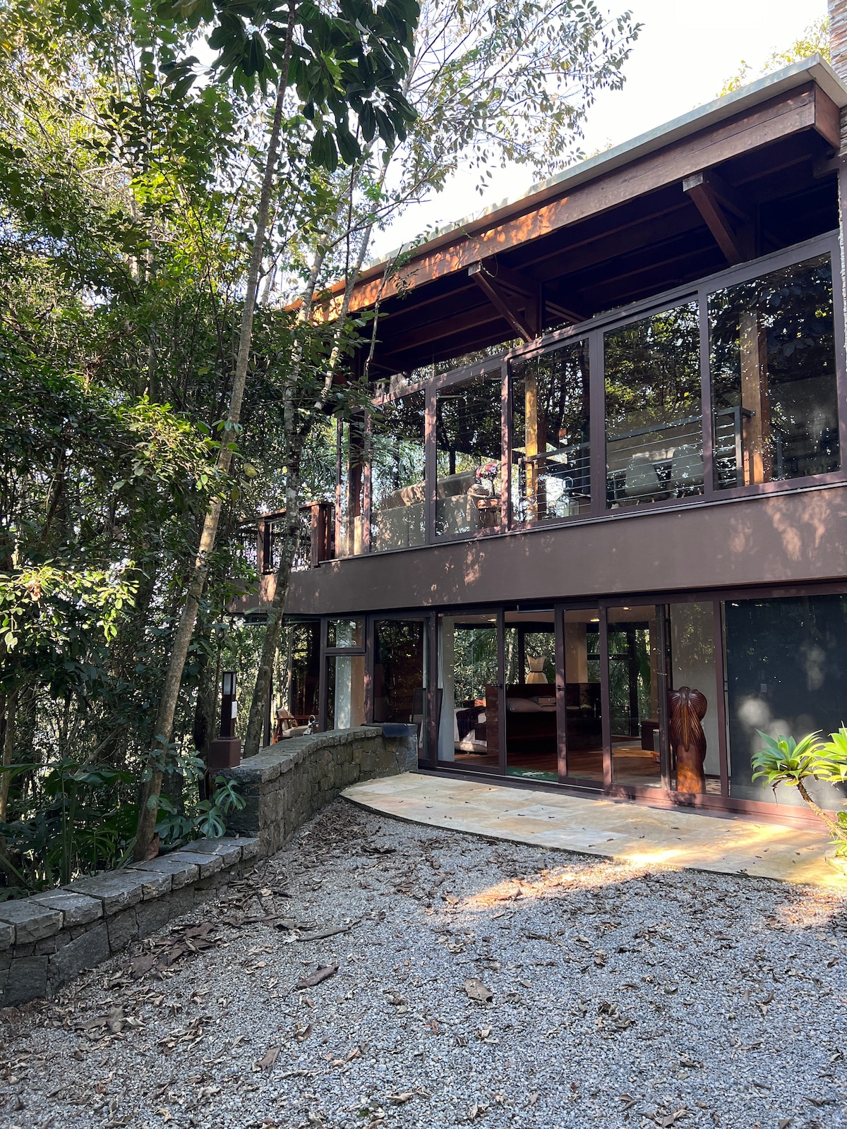 A modern, two-story glass house is surrounded by lush greenery. The exterior features large windows and wooden beams, blending seamlessly with the natural landscape. A stone pathway leads to the entrance, enhancing the house's connection to the tranquil environment.