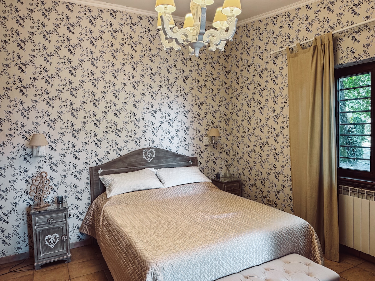 A spacious bedroom features a large bed with a soft beige coverlet, framed by floral-patterned walls. Nightstands with decorative lamps are placed on either side of the bed. A stylish chandelier provides overhead lighting, and a window allows natural light to filter in through light curtains.