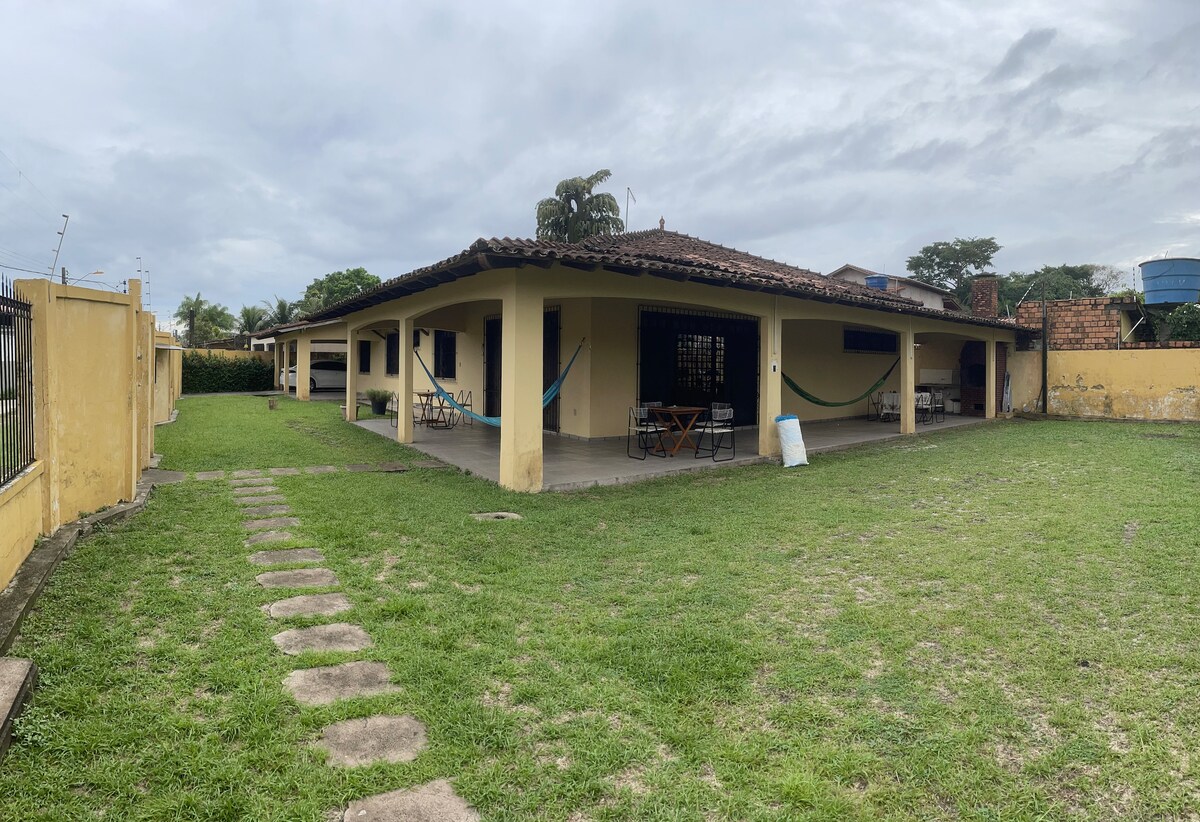 A spacious outdoor area surrounds a single-story house, featuring a well-maintained lawn and pathways. The property includes shaded seating areas and an adjoining covered patio for relaxation. A water tank is visible in the background, along with greenery and trees, enhancing the serene atmosphere.