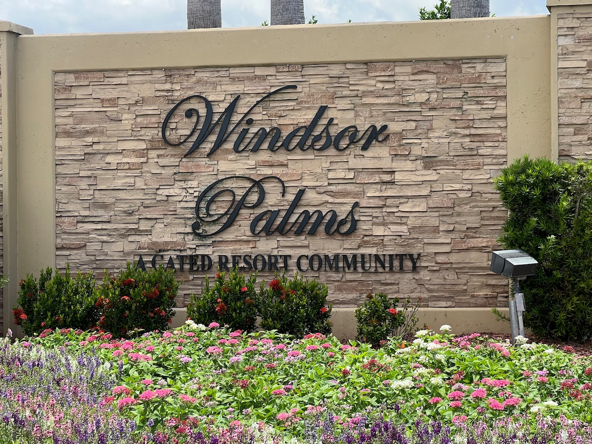 The entrance to Windsor Palms Resort is marked by a stone sign displaying the name in elegant lettering. Surrounding the sign are vibrant flower beds filled with colorful blooms, enhancing the welcoming appearance of the resort community.