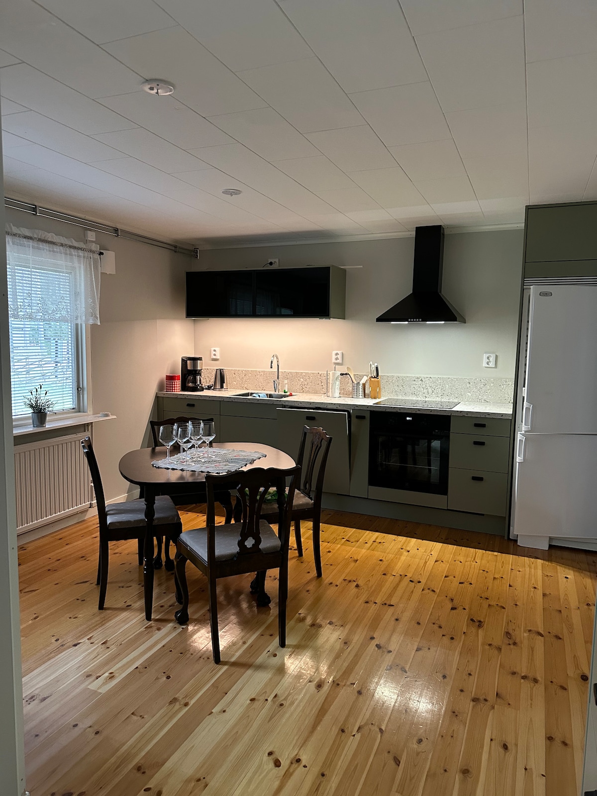 A modern kitchen is presented, featuring a compact dining area with a round table and four chairs. The kitchen includes a countertop, stove, oven, and refrigerator. Natural light enters through a window, reflecting off the polished wooden floor.