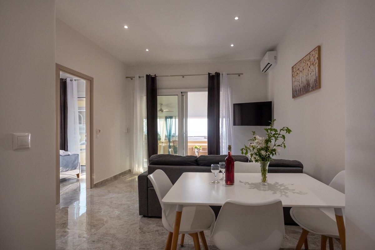 A modern living area features a light-colored table surrounded by four chairs, with a decorative vase of greenery at its center. Soft gray sofas are positioned nearby, and a small television is mounted on the wall. Large windows allow natural light to fill the space.