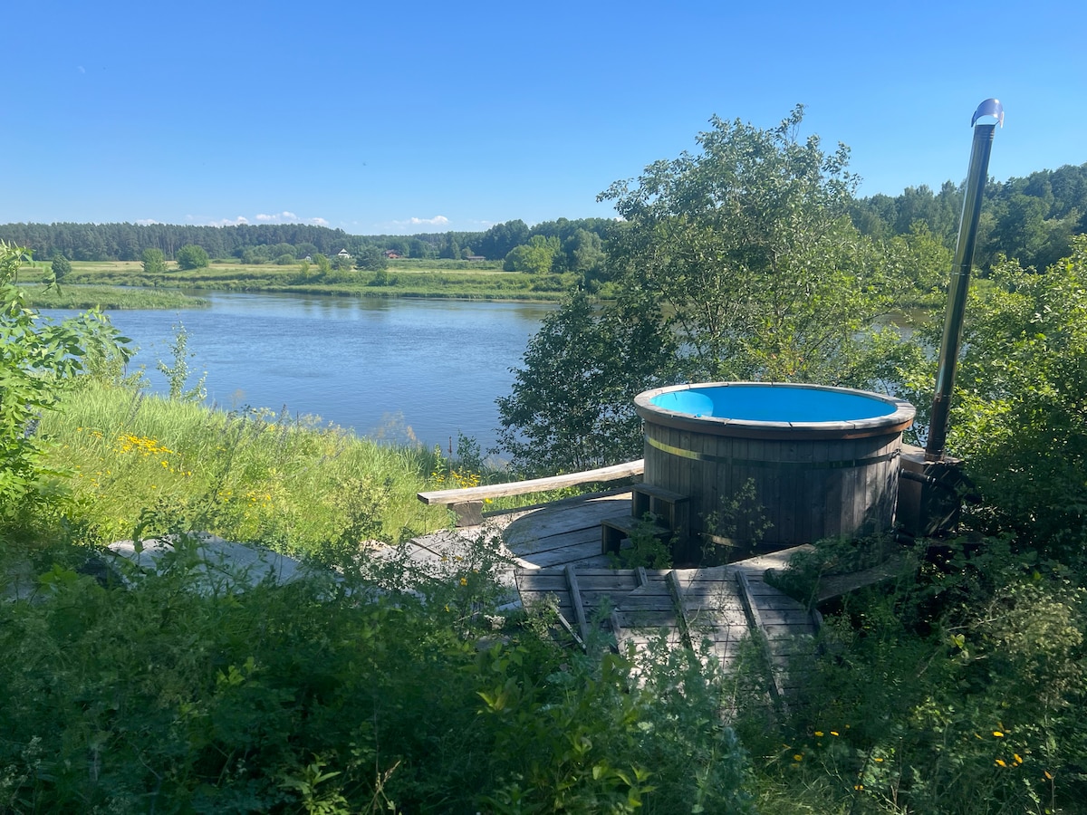 A wooden hot tub is situated on a wooden deck overlooking a calm river. Lush greenery surrounds the area, and distant trees are visible on the opposite bank. The clear blue sky reflects on the water, creating a serene outdoor environment.