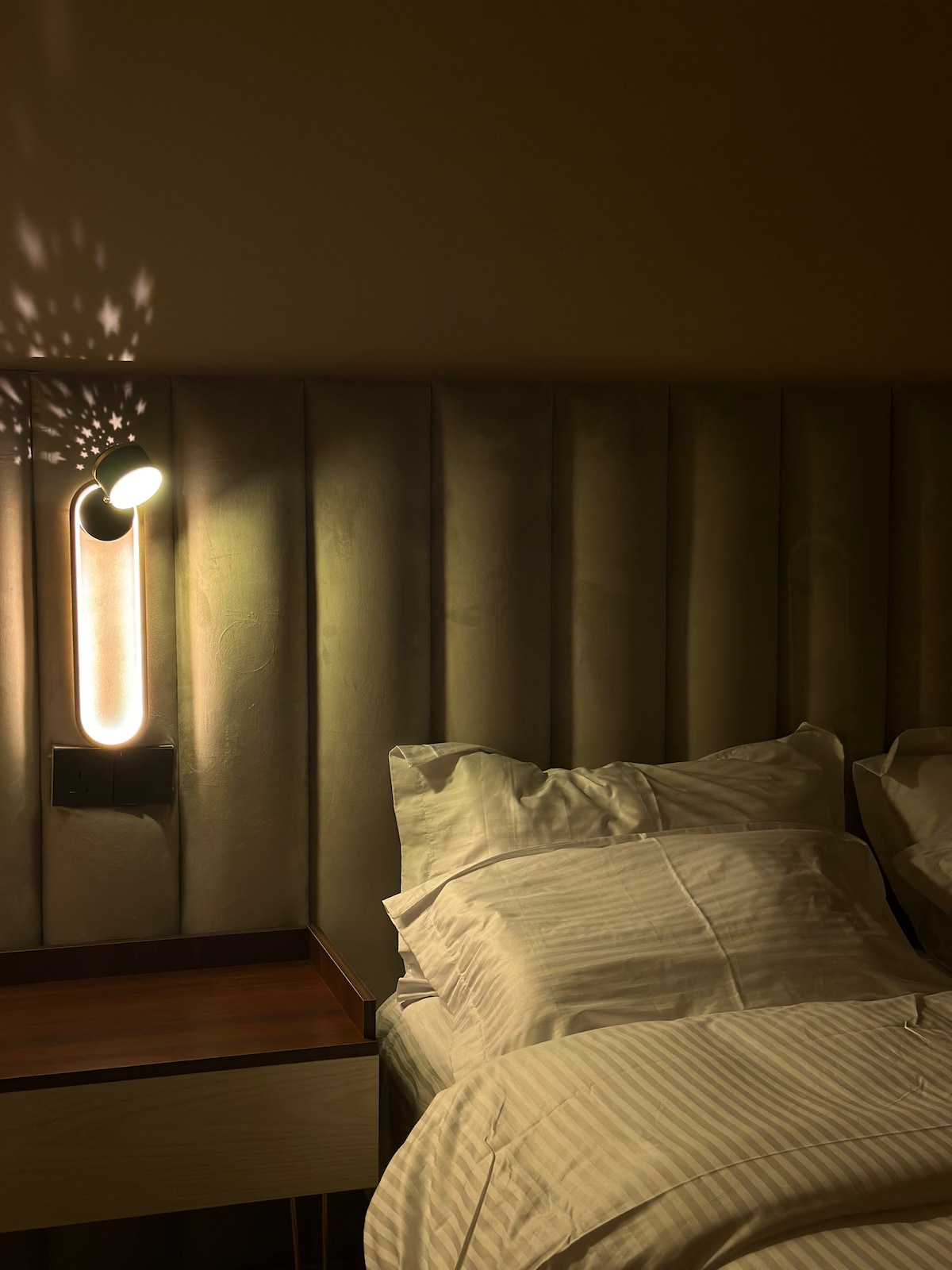 A cozy sleeping area features soft bedding arranged on a spacious bed, with a subtle headboard design that adds texture. A wall-mounted light illuminates the space, casting gentle shadows, while a nightstand holds a modern lamp, enhancing the room's calming ambiance.