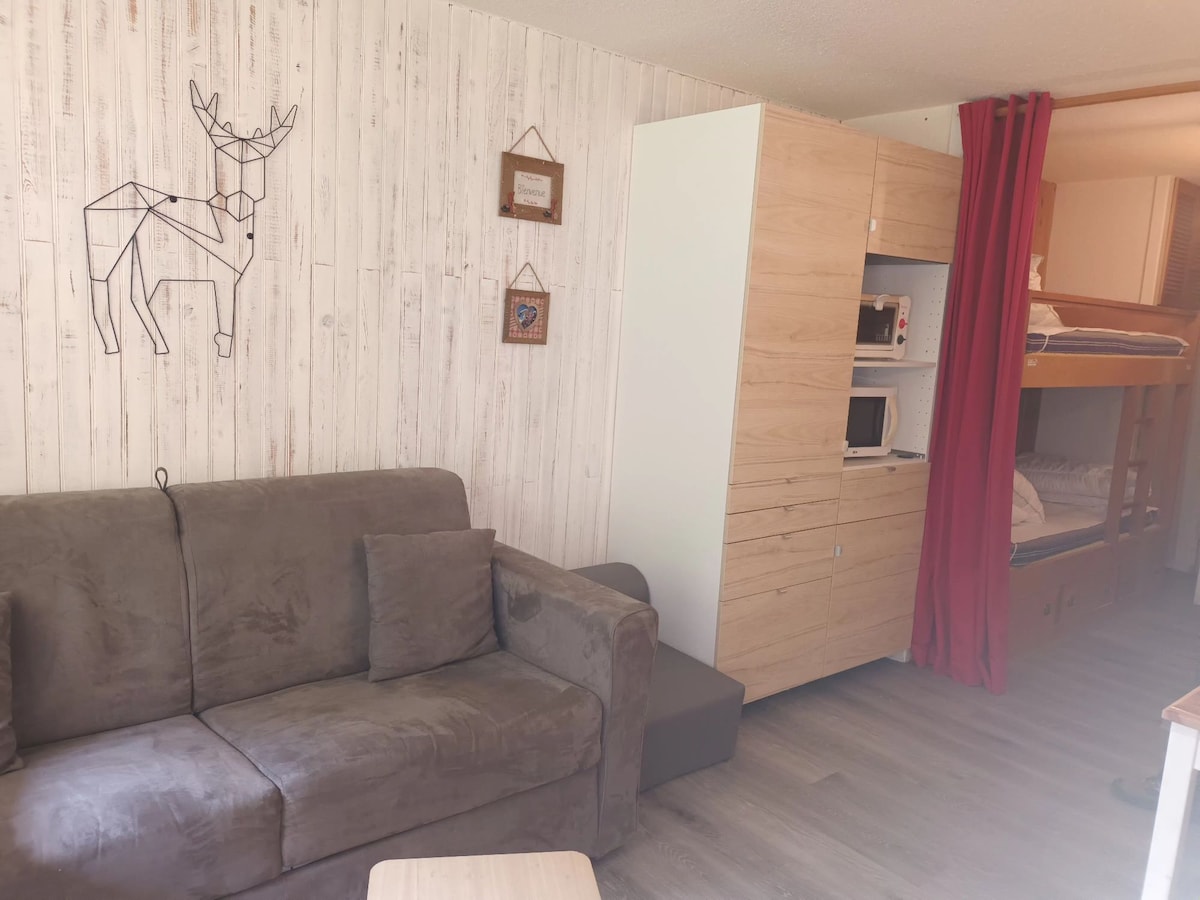 A living area features a comfortable gray sofa with decorative pillows against a wooden wall. A minimalist storage unit, including a microwave, is positioned nearby. To the side, a partial view of the bunk beds is visible, separated by a red curtain.